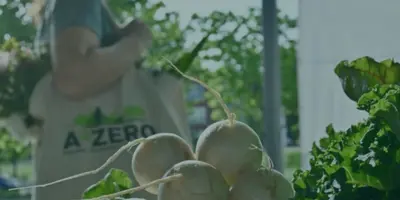 A person holding a reusable bag with the A2ZERO logo on it stands behind a farmers market table with piles of turnips and radishes on top.