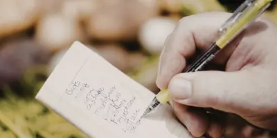 Person writes a grocery list on a small notepad in front of a food display.