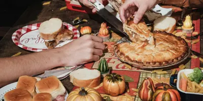 Three people reach into the center of a table filled with food, including pies, squash, and biscuits.