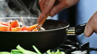 Two hands prepare food in a steaming pan behind green leafy vegetables.